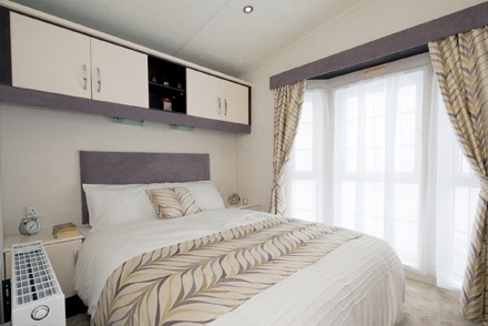 A static caravan’s double bedroom, there are cupboards above the bed and a big window behind. The bed is made with attractive modern bed linen.