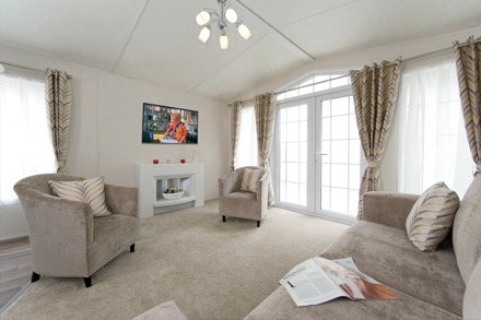 Beige carpets, matching comfy furniture, white walls, white ceiling and classy full length brown drapes compliment this open plan lounge. The room is flooded with light from the full glass French doors to the right. It’s amazing it’s a static caravan not a luxury villa!!