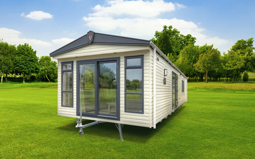 The end view of a brand new static caravan, it’s positioned on mowed grass. It is cream with grey trims. The tow bar is showing indicating it’s going to be moved.