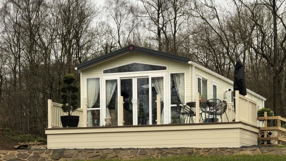 This new static caravan, on the edge of the Wyre forest, has a huge array of windows to the nose end and around the sides. It has tall trees behind and sits on the top of a grassey slope above the quiet fishing pool.